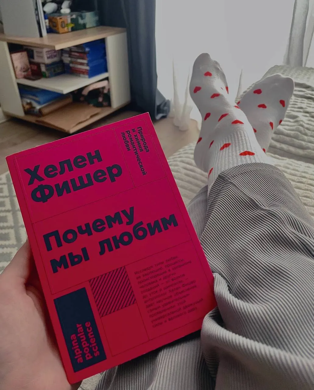 Книга Хелена Фишера “Почему мы любим” в руке на фоне мягкой и светлой кровати